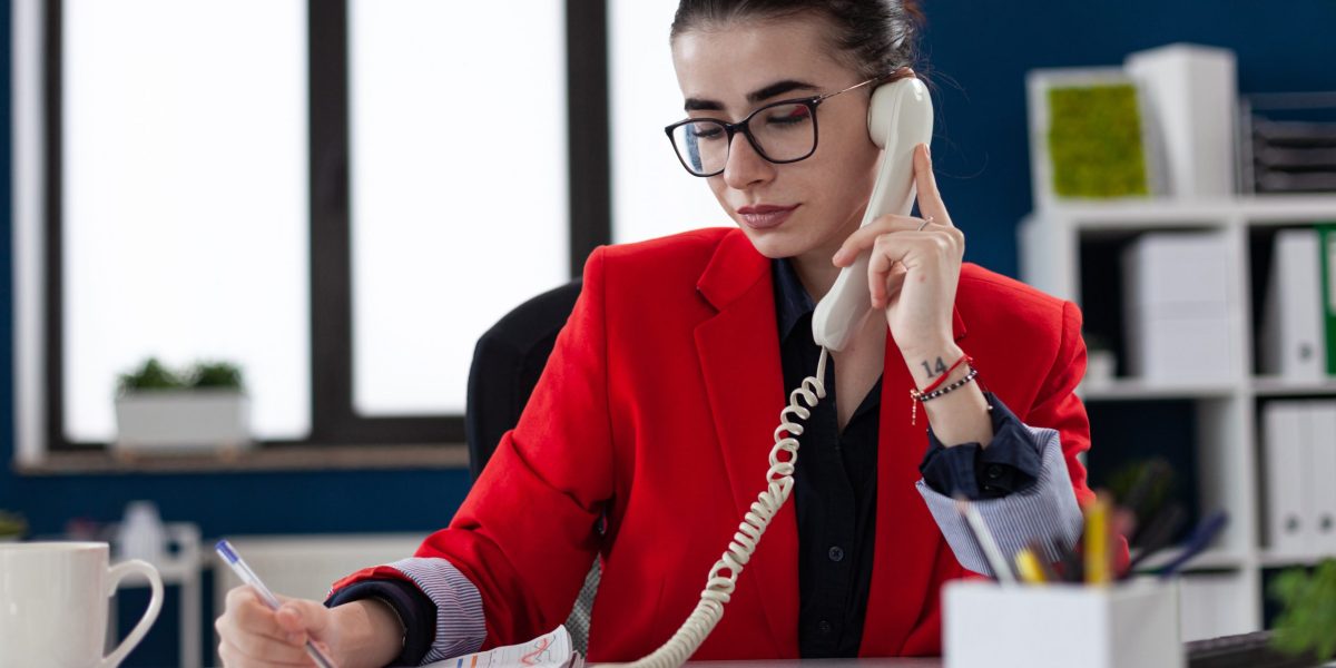 Businesswoman taking notes on clipboard sitting at desk in corporate office, while taking with adviser about financial expertise. Busy manager having a conversation about deadline.